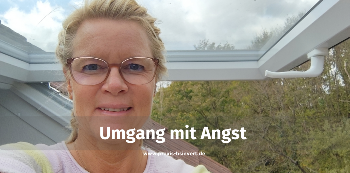 Selbstporträt einer lächelnden Frau mit blonden, geflochtenen Zöpfen und Brille an einem geöffneten Dachfenster, im Bild der Schriftzug „Umgang mit Angst“