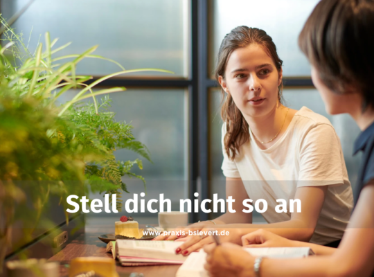 Zwei Frauen sitzen sich an einem Tisch im Café gegenüber und sprechen miteinander, eine Pflanze im Vordergrund, im Bild der Text „Stell dich nicht so an"